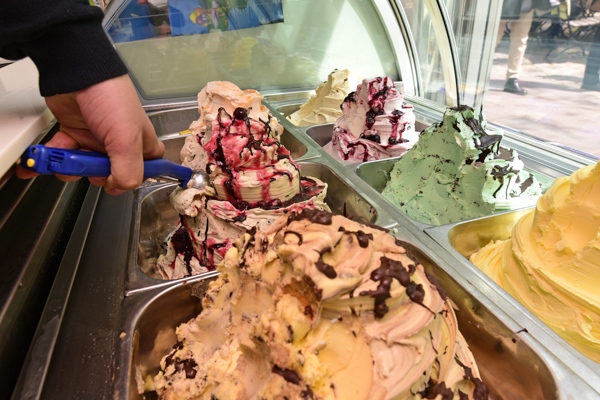 Eine Person schöpft bunte Eissorten mit Schokolade und Soße aus einer Eistheke