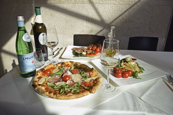 Ein gedeckter Tisch mit Pizza, Pasta, Wein und Wasser in hellem Sonnenlicht