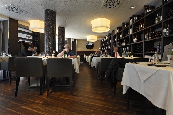 Zwei Männer mit ernster Miene sitzen an gedeckten Tischen in einem modernen Restaurant mit dunklen Holzböden und einer auffälligen Wand voller Weinflaschen
