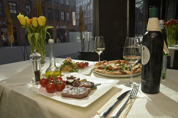 Ein festlich gedeckter Tisch mit Pizza, Steak, frischem Gemüse und Wein in hellem Licht
