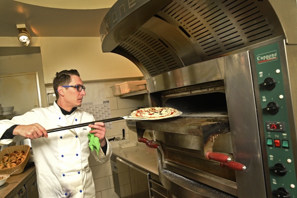 Ein Mann mit Brille und weißer Kochjacke schiebt konzentriert eine Pizza in den Ofen