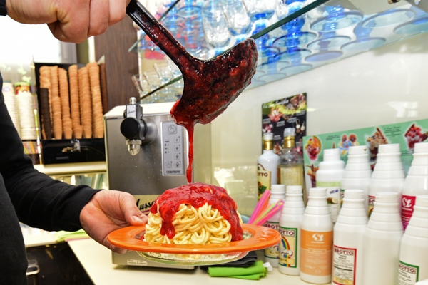 Eine Person gießt rote Soße über ein Spaghetti-Eis auf einem orangefarbenen Teller